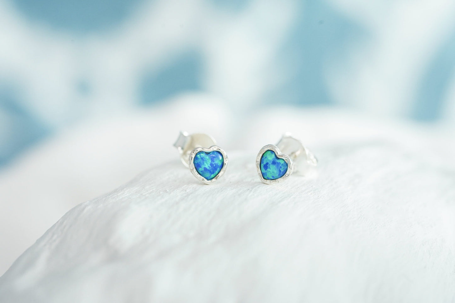 Small Sterling Silver Opalite Heart Studs on a white surface with a blurred blue background