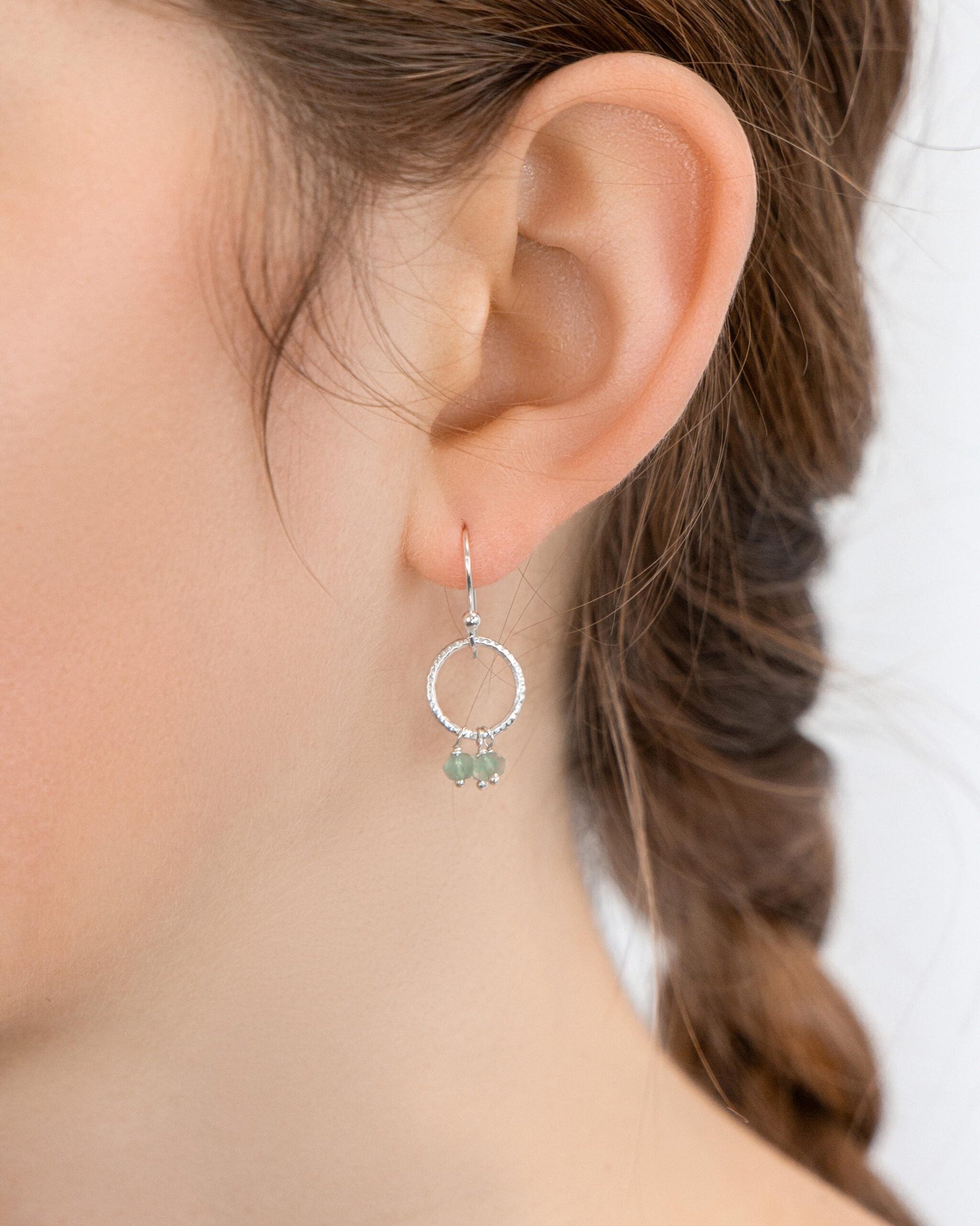 Close-up of an ear wearing a Joli Beau Sterling Silver Drop Hoop Aqua Marine Earrings against a neutral background