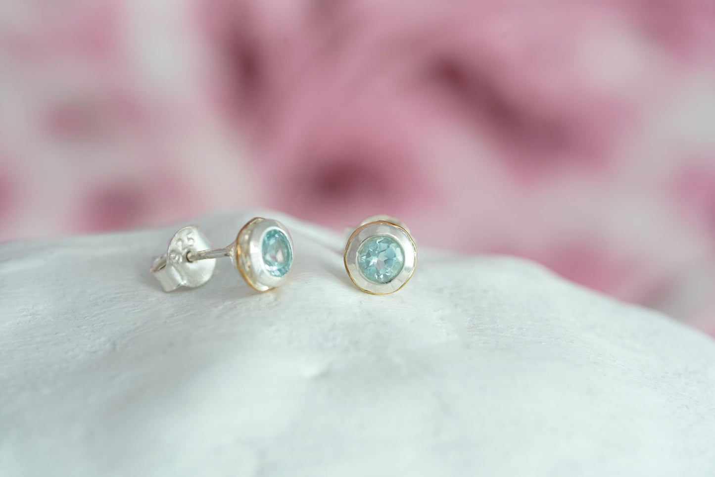 Pair of Sterling Silver Topaz With Gold Detail Studs on a white surface with a blurred pink background