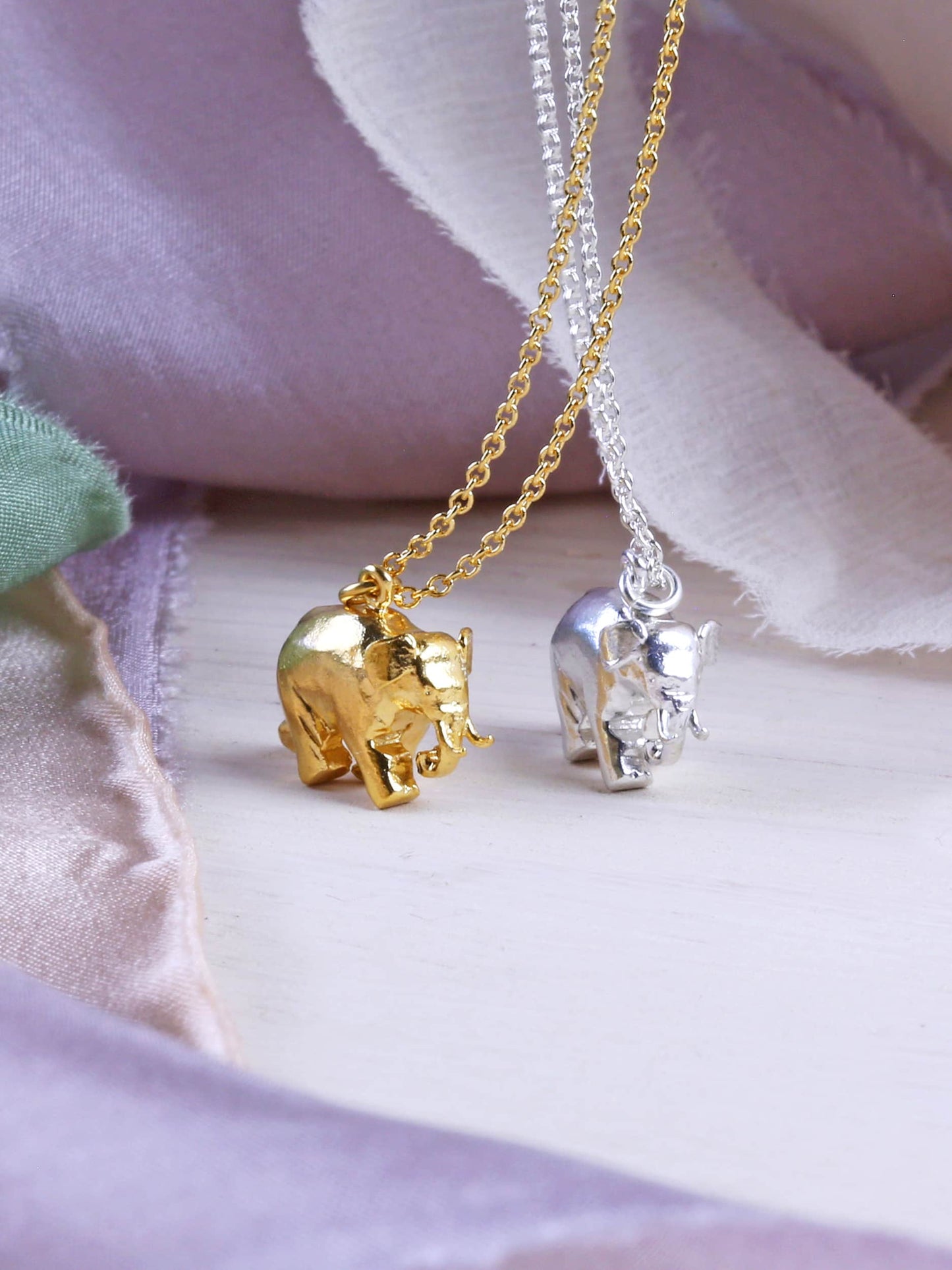 Gold and silver elephant pendants on chains against a soft fabric background