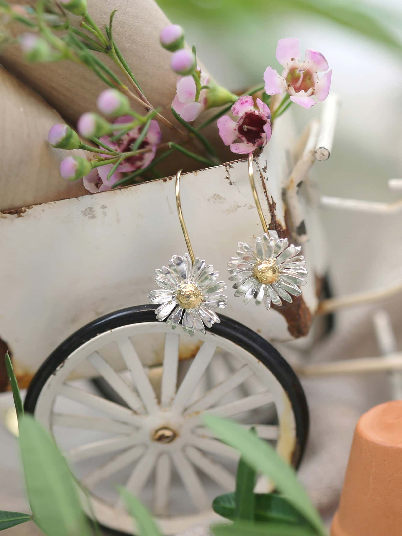 Alex Monroe 22 carat Gold Vermeil & Sterling Silver Big Daisy Hook Earrings with a decorative background featuring a bicycle wheel and flowers.