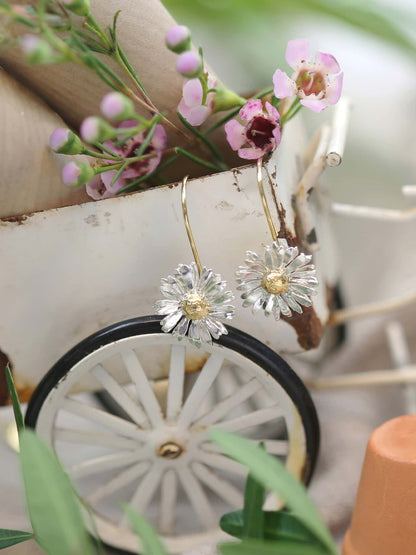 Alex Monroe 22 carat Gold Vermeil & Sterling Silver Big Daisy Hook Earrings with a decorative background featuring a bicycle wheel and flowers.