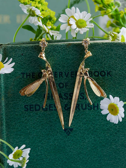 Alex Monroe Statement 18 carat Vermeil Damsel Fly & Grassblade Green Amethyst Drop Earrings on a green book with white flowers