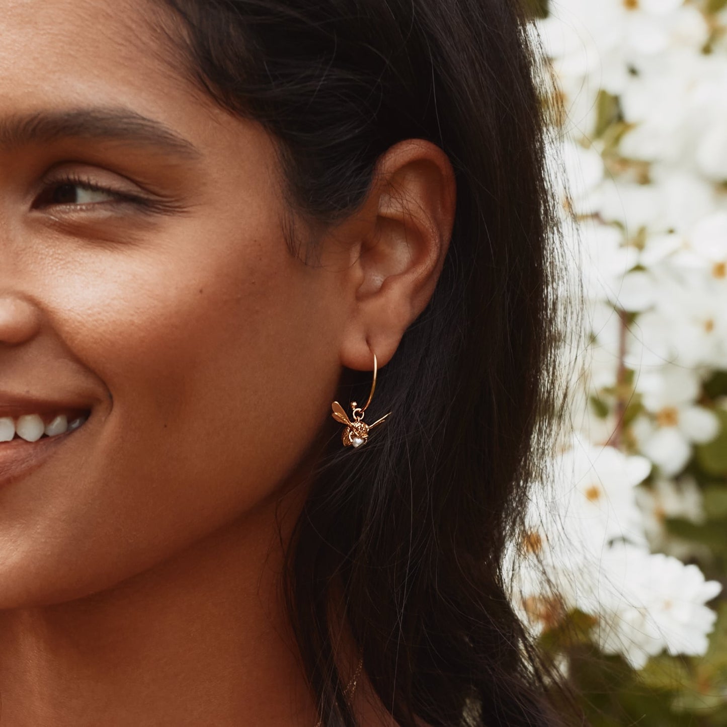 Close-up of a woman wearing a gold bee-shaped earring with a blurred floral background