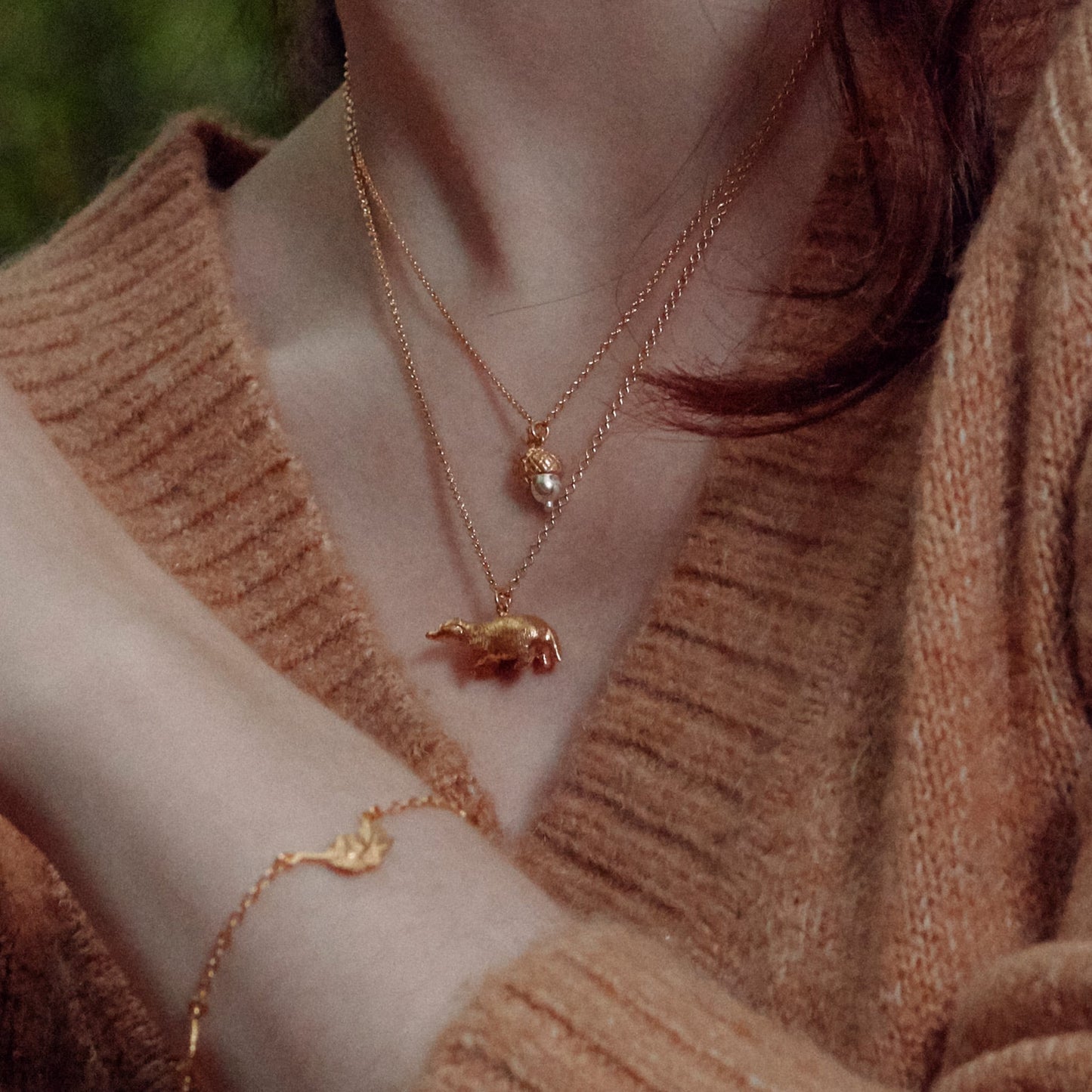 Close-up of a person wearing an Alex Monroe 22 carat Yellow Gold Vermeil Foraging Badger necklace and bracelet with a soft brown background