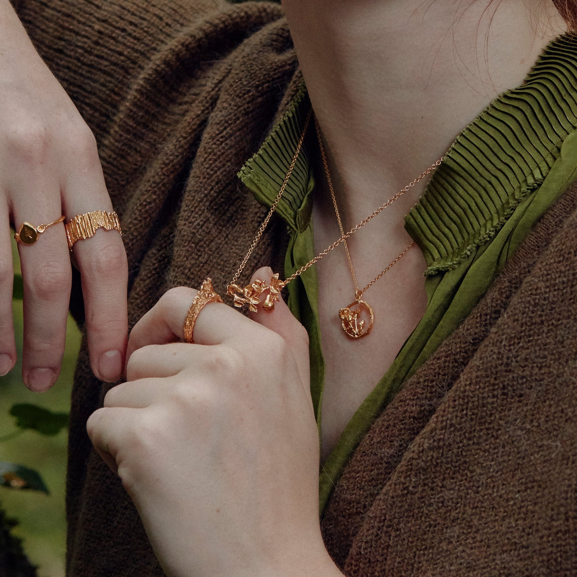 Close-up of hands wearing gold rings and necklaces, with a blurred natural background.