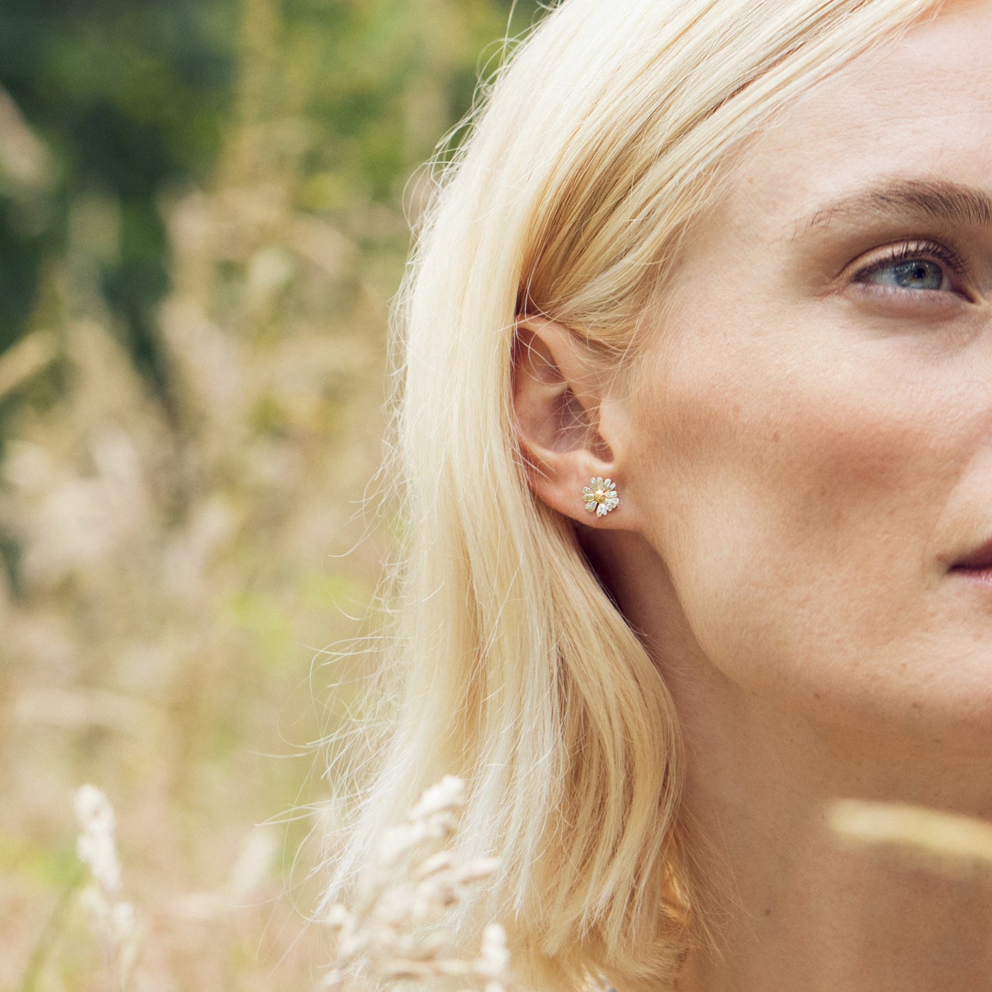 Close-up of a woman wearing Alex Monroe Mixed Silver & Gold Vermeil Little Daisy Stud Earrings with a blurred natural background