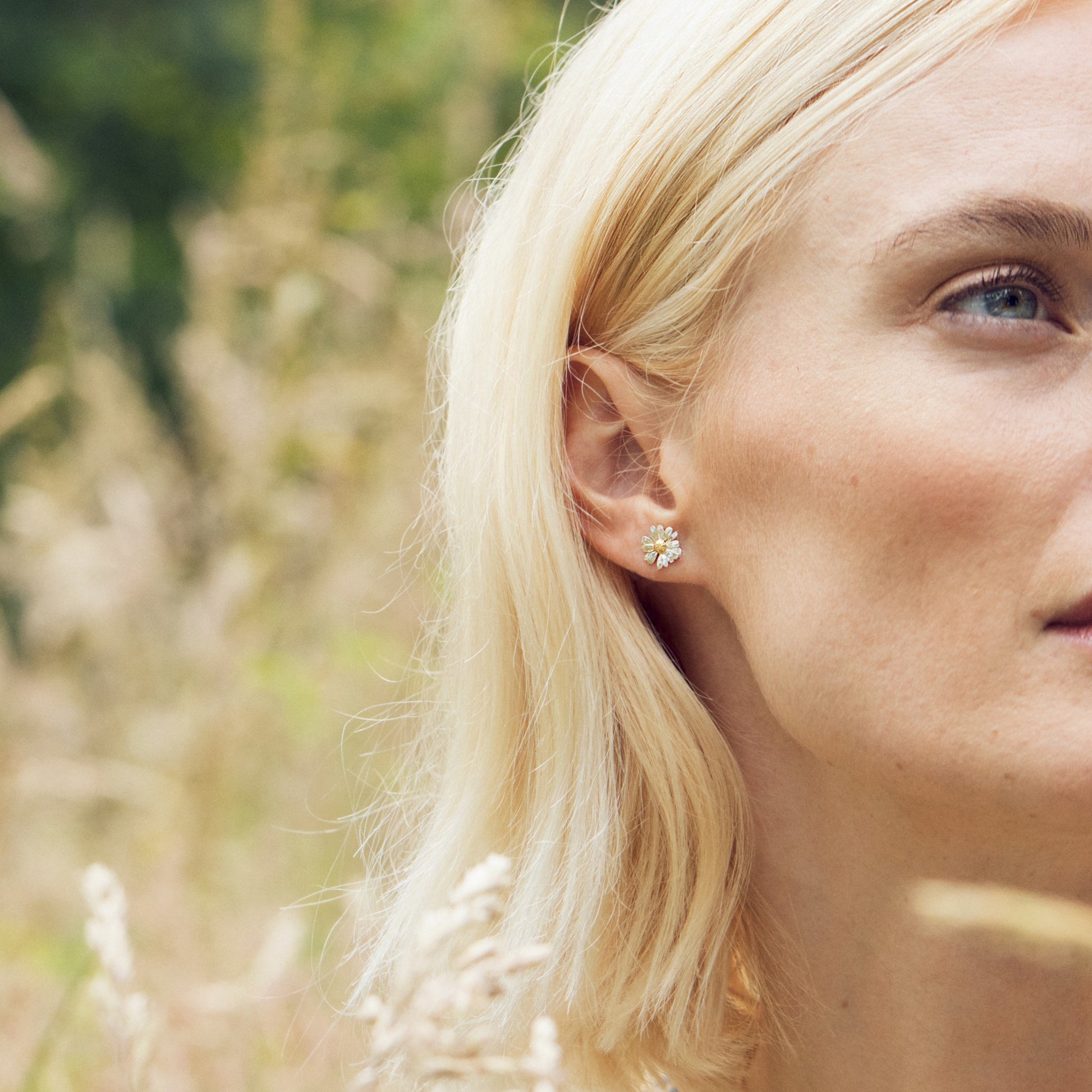 Close-up of a woman wearing Alex Monroe Mixed Silver & Gold Vermeil Little Daisy Stud Earrings with a blurred natural background