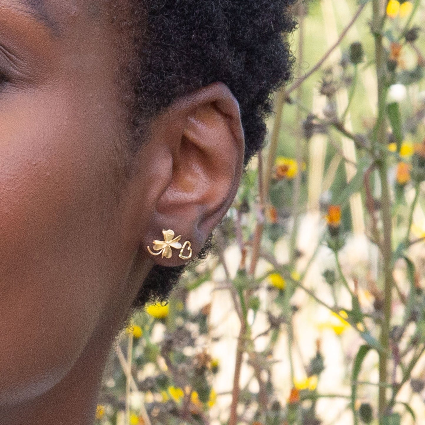 Close-up of a person's ear with a Alex Monroe 22 carat Yellow Gold Vermeil Lucky Clover Stud Earring against a natural background