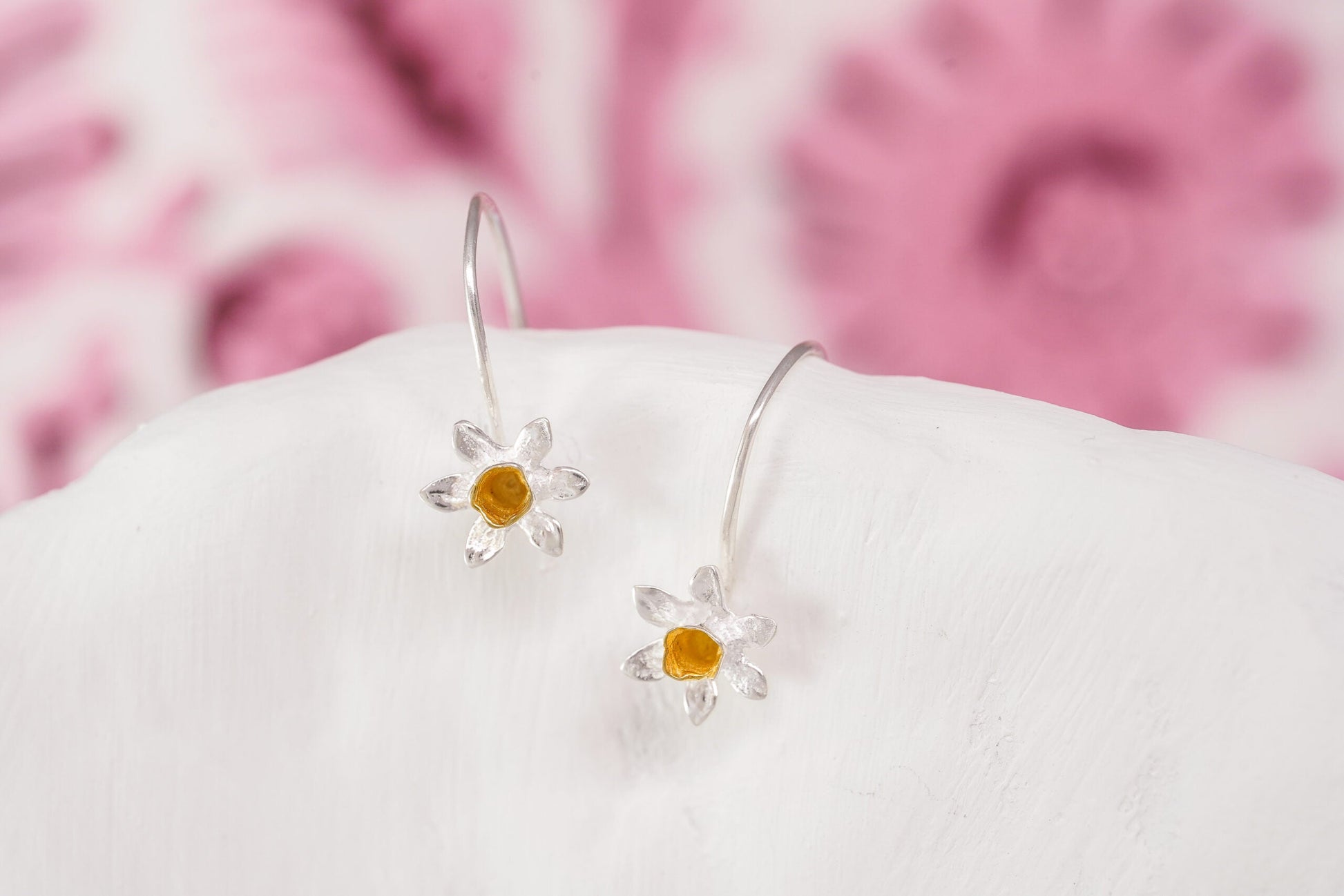 Silver daisy earrings on a white surface with a pink floral background