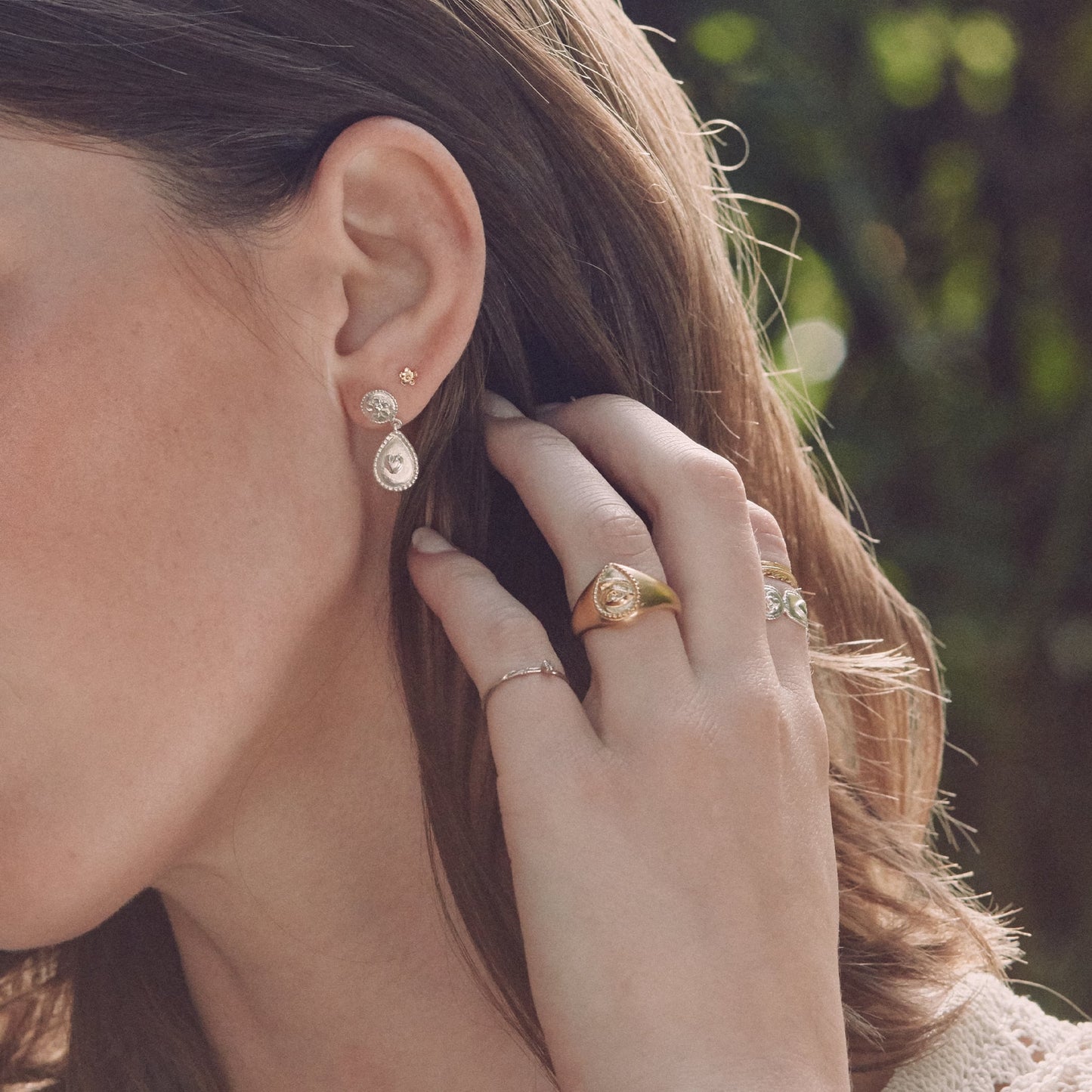 Close-up of a woman's ear with Alex Monroe Sterling Silver Love of Nature Heart & Flower Drop Earrings and hand with rings, blurred green background