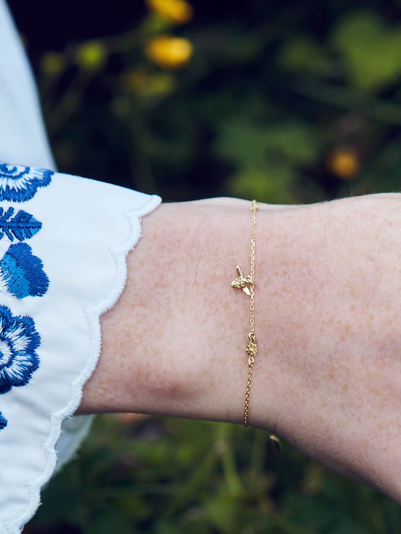 Alex Monroe 18 carat Yellow Solid Gold The Beekeeper Floral Chain Bracelet on a wrist with a blurred natural background