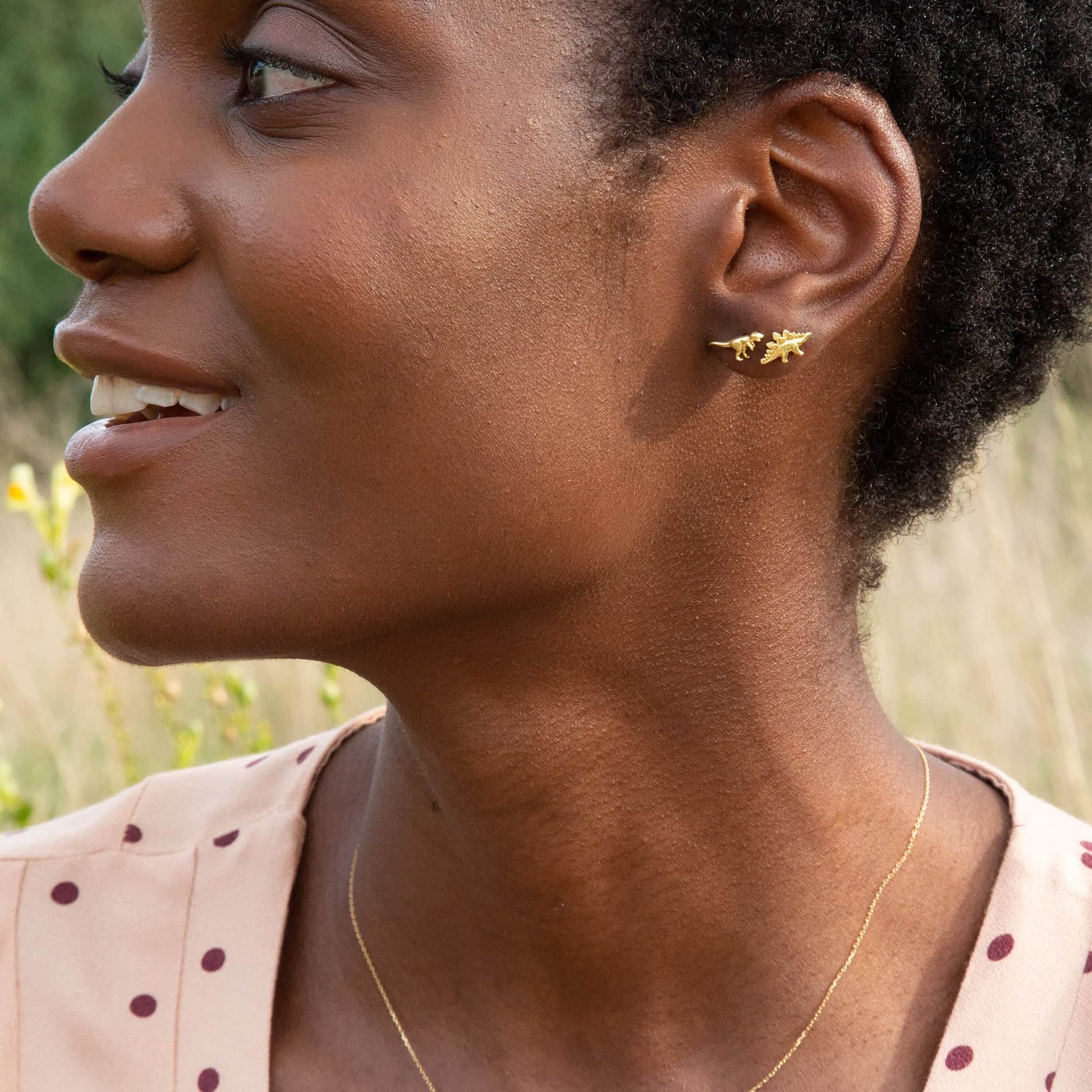 Woman wearing a Alex Monroe 18 carat Yellow Solid Gold Teeny Tiny T-Rex Single Stud Earring and necklace outdoors