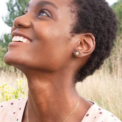 Woman wearing Alex Monroe 18 carat Yellow Solid Gold Teeny Tiny Diamond Daisy Stud Earrings and a necklace in a natural setting