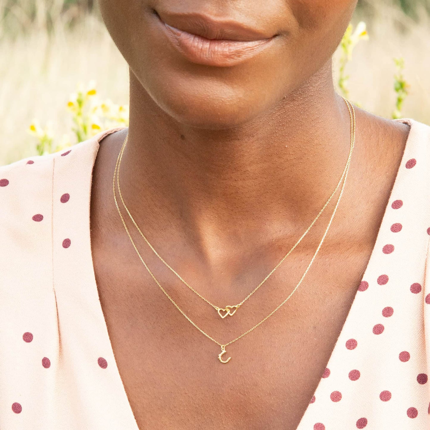 Woman wearing a gold necklace with heart and crescent pendants against a blurred natural background.